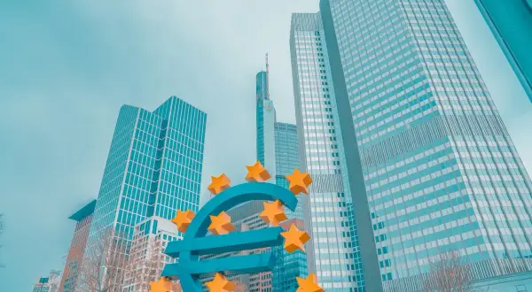 Duży, niebieski znak euro z żółtymi gwiazdami, na tle nowoczesnych wieżowców.