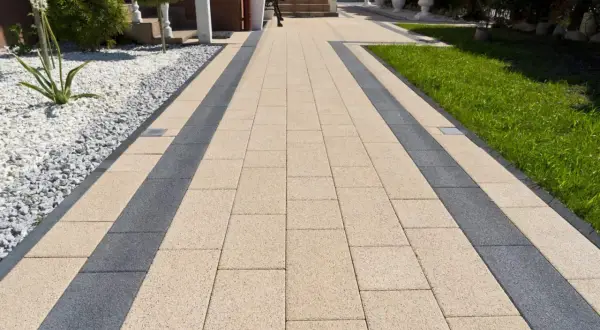 Eleganckie ścieżki z kostki brukowej w beżowo-grafitowym wzorze prowadzą do domu. Otoczone zielenią i białym żwirem.