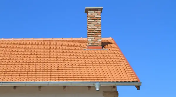 Ceglany komin na pomarańczowym dachu pod błękitnym niebem.