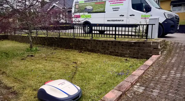 En robotgräsklippare från Honda står på en gräsmatta. En vit servicebil med texten "TrädgårdSmart" och "gardena blinkar blått" står parkerad bakom ett staket.