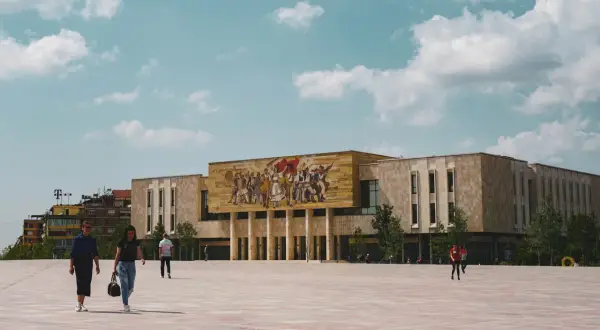 Plac w centrum Tirany w Albanii, z monumentalnym budynkiem ozdobionym mozaiką i ludźmi spacerującymi po rozległym, wybrukowanym placu.