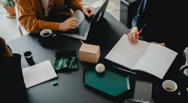 Två personer vid ett bord, en med laptop, den andra skriver i en bok. Kaffe och anteckningsmaterial. De är bi utvecklare som samarbetar.