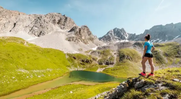 Une femme admire un lac de montagne lors d'une randonnée à Courchevel. Paysage alpin spectaculaire avec des montagnes rocheuses et des prairies verdoyantes.