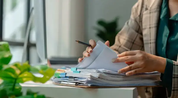 Pracownik przegląda dokumenty, marząc o urlopie dla poratowania zdrowia. Stos papierów czeka na biurku.