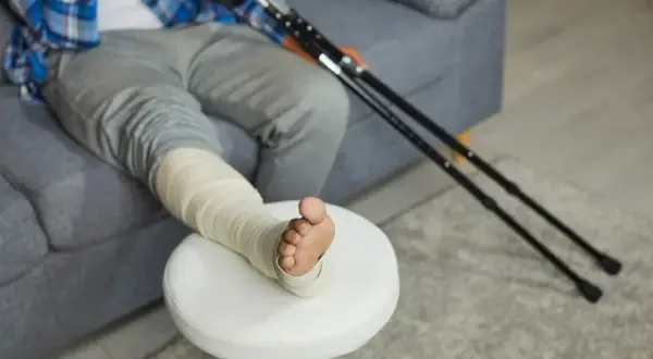 Osoba z zabandażowaną nogą i kulami łokciowymi siedzi na kanapie.