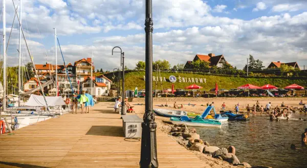 Słoneczne plaże na Mazurach tętnią życiem. Ludzie odpoczywają na piasku, kąpią się w jeziorze i pływają łódkami.