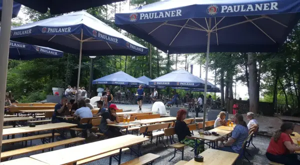 Ein sonniger Tag im Paulaner Biergarten. Gäste sitzen an Holztischen unter großen Sonnenschirmen und genießen die Atmosphäre.
