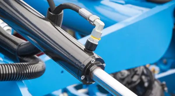 Części do hydroforu: czarny siłownik hydrauliczny z metalowym złączem i wężem, na tle niebieskiej maszyny.