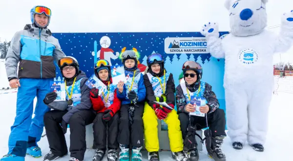 Uśmiechnięci młodzi narciarze z dyplomami i medalami po zajęciach w szkole narciarskiej Koziniec. Towarzyszy im instruktor i biały miś.
