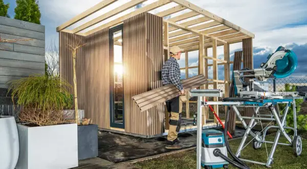 Costruzione di una casetta in legno: un uomo porta pannelli per completare la struttura, con sega troncatrice e aspiratore pronti all'uso.