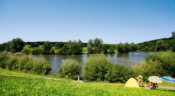 Sonniger Tag am Aichstruter Stausee. Menschen planschen im Wasser, baden und entspannen sich am Ufer unter bunten Sonnenschirmen.