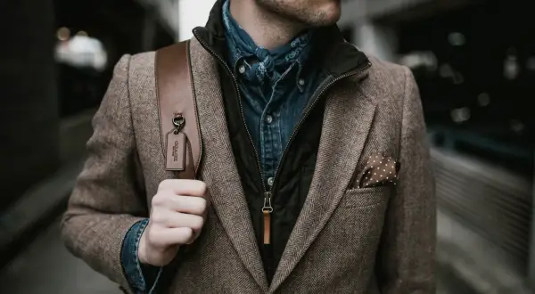 Mann im Tweed-Sakko mit Jeanshemd und Halstuch, perfekt für den party chic dress code male. Braune Ledertasche mit Coach-Anhänger.