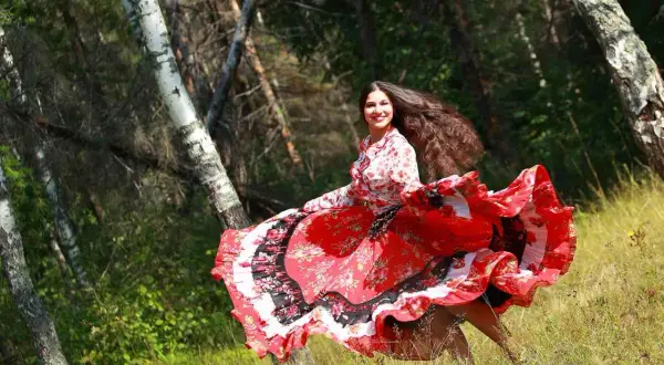 Una giovane donna con lunghi capelli scuri, vestita con un abito rosso svolazzante, danza in un bosco. Sembra una delle giostrai Sinti, piena di gioia e vitalità.