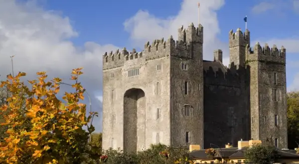 El imponente castillo de Bunratty se alza bajo un cielo azul con nubes, rodeado de follaje otoñal.