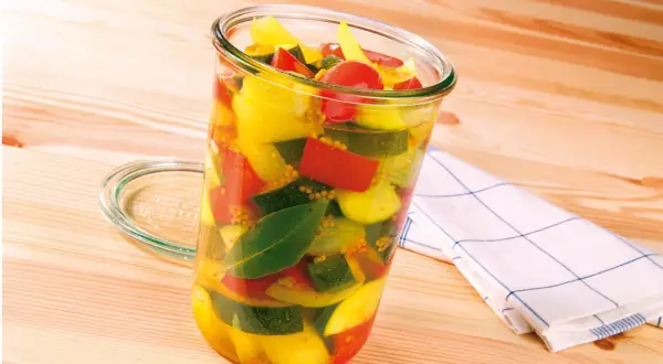 Ein Glas mit bunter Mischung aus Zucchini, Paprika und Tomaten, bereit zum Zucchini einkochen. Senfkörner und Lorbeerblätter sind ebenfalls sichtbar.
