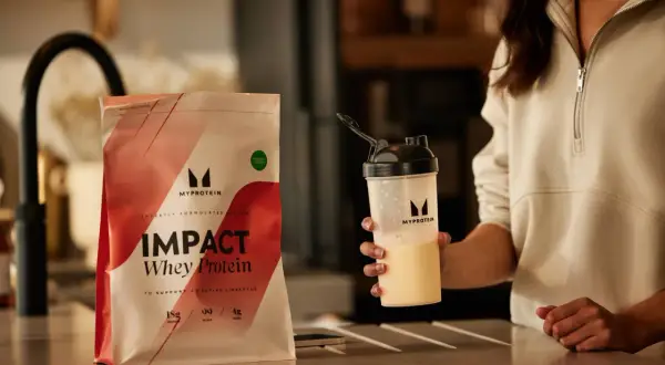 Eine Person hält einen Shaker mit einem cremigen Getränk. Daneben liegt eine Packung Myprotein Impact Whey Protein. Perfekt für den proteinshake vor dem schlafen.