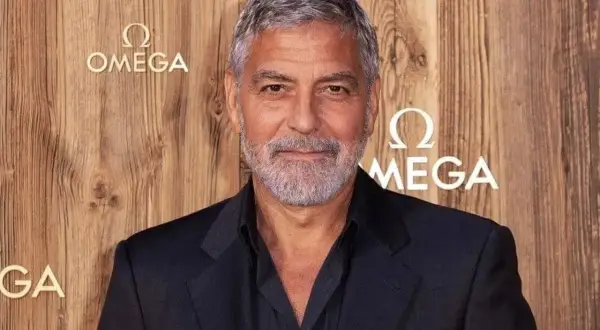 Un homme avec des cheveux gris courts et une barbe, portant un costume sombre, pose devant un mur en bois avec le logo Omega.