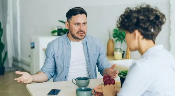 Mężczyzna z wyrzutem mówi do kobiety, która zdaje się ignorować narcyza, skupiając się na jabłkach.