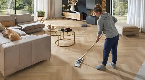 Frau wischt den hellen Designboden im Wohnzimmer. Ein Saugroboter ist im Hintergrund zu sehen.