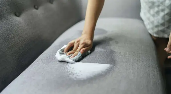 A hand with a soapy cloth gently cleans a grey couch cushion, showing how to wash couch cushion covers for a fresh look.