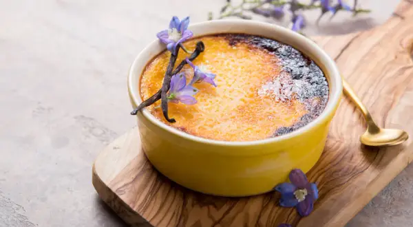 Creme brulee rezept: goldbraune Kruste, Vanilleschoten und blaue Blüten zieren das Dessert auf einem Holzbrett.