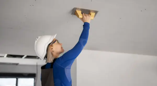 Kobieta w kasku i okularach przykleja styropian do sufitu za pomocą kielni.