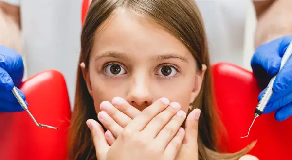 Przerażona dziewczynka zakrywa usta dłońmi, gdy dentysta przygotowuje się do podania znieczulenia. Wokół niej widać czerwony fotel dentystyczny i narzędzia.