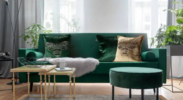 Zielona kanapa z ozdobnymi poduszkami z motywem pszczoły, złotymi stolikami i pufą.