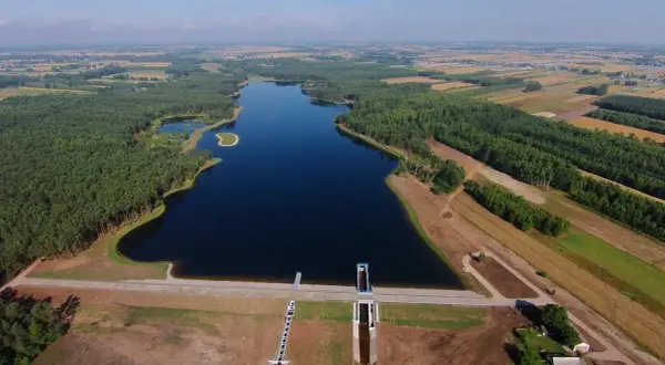 Widok z lotu ptaka na zalew Jagodno otoczony lasem i polami. Woda lśni w słońcu, a tama z budowlami hydrotechnicznymi stanowi imponującą konstrukcję.