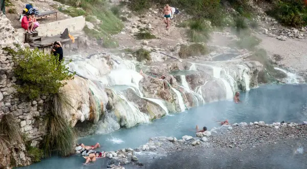 Persone si rilassano nelle acque fumanti delle terme di Petriolo, tra rocce calcaree e vegetazione.