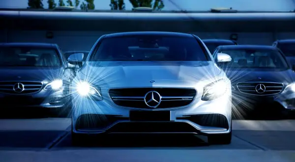 Biała hybryda plug-in Mercedes-Benz z włączonymi światłami, otoczona innymi ciemnymi samochodami tej marki.
