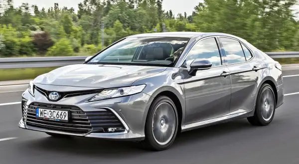 Srebrna Toyota Camry 2023 jedzie po drodze, otoczona zielenią drzew.
