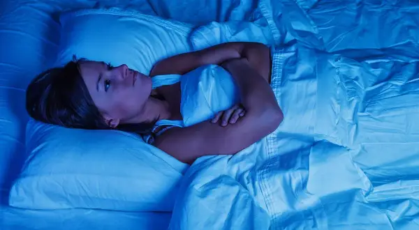 Frau liegt wach im Bett, Arme verschränkt, die Augen weit aufgerissen. Deutliche Anzeichen von Schlafmangel und dessen Folgen.