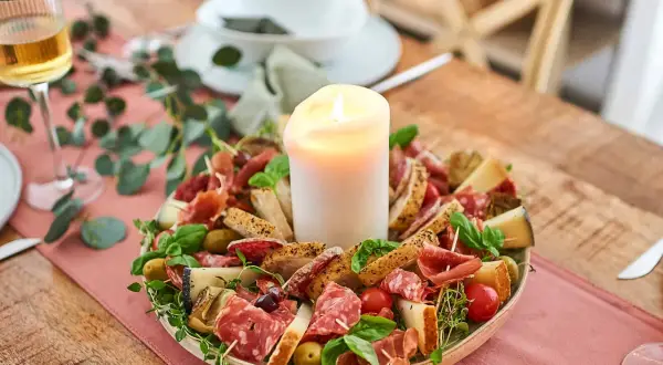 Antipasti selber machen: Eine festliche Platte mit Wurst, Käse, Brot, Oliven und Tomaten, umgeben von grünen Zweigen, mit einer Kerze in der Mitte.
