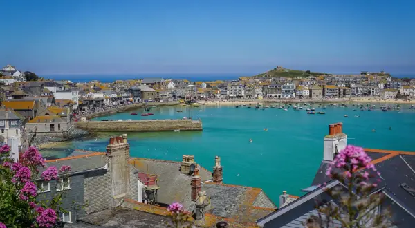 En solig vy över St Ives med turkost vatten, båtar och hus. Blommor i förgrunden ger en somrig känsla.