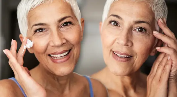 Zwei Frauen mit kurzen weißen Haaren lächeln. Eine trägt Creme auf ihre Wange auf, um trockene Haut im Alter zu bekämpfen.