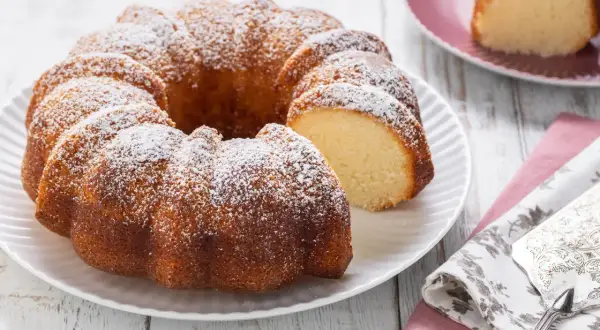Ciambellone senza uova, soffice e dorato, spolverato di zucchero a velo. Una fetta è già stata tagliata, invitando ad assaggiarlo.