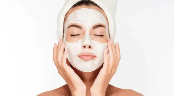 Une femme avec une serviette sur la tête applique un masque blanc sur son visage, espérant atténuer les marques de masque cicatrice acné.