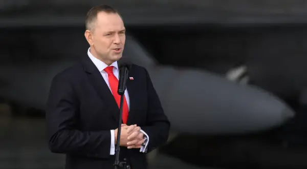 Mężczyzna w garniturze przemawia przy mikrofonie, w tle samolot. Mówi o reparacje wojenne od niemiec.