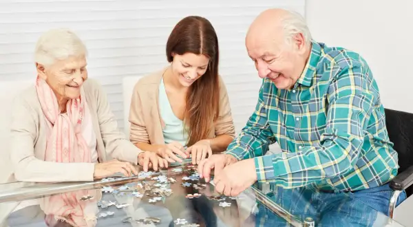 Trois personnes, deux aînées et une jeune femme, s'amusent à assembler un puzzle, une activité stimulante pour les jeux de mémoire.