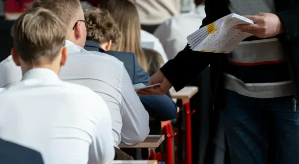 Nauczyciel rozdaje arkusze egzaminacyjne uczniom siedzącym w ławkach.
