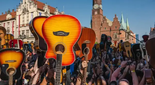 Tłum ludzi z gitarami wzniesionymi ku górze, świętujący gitarowy rekord świata na tle zabytkowej architektury.