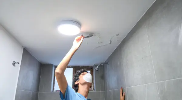 Mężczyzna w masce usuwa łopatką łuszczącą się farbę z sufitu w łazience.
