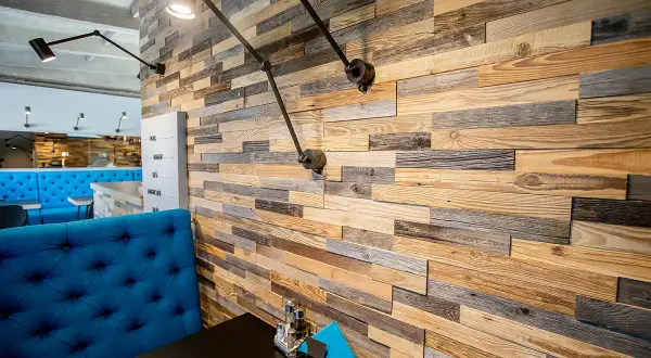 Mur en bois brut, aux teintes variées, évoquant l'essence de bois authentique. Des appliques industrielles éclairent ce décor chaleureux.