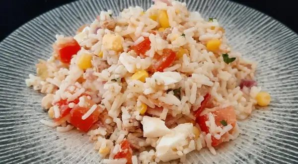 Une généreuse portion de salade de riz thon, garnie de dés de tomates, de maïs et d'œufs durs, sur une assiette texturée.