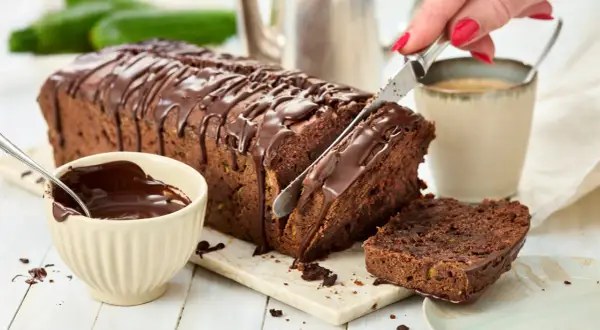 Ein saftiger Zucchini Schoko Kuchen wird angeschnitten. Daneben eine Schale mit Schokoladenglasur und eine Tasse Kaffee.