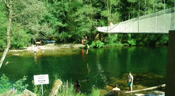 Puente colgante sobre río verde con gente bañándose y disfrutando del paisaje.
