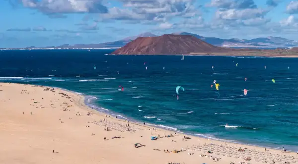 Fuerteventura, gdzie najlepiej? Plaża z kitesurferami i turystami, w tle wulkan i góry. Idealne miejsce na wakacje.