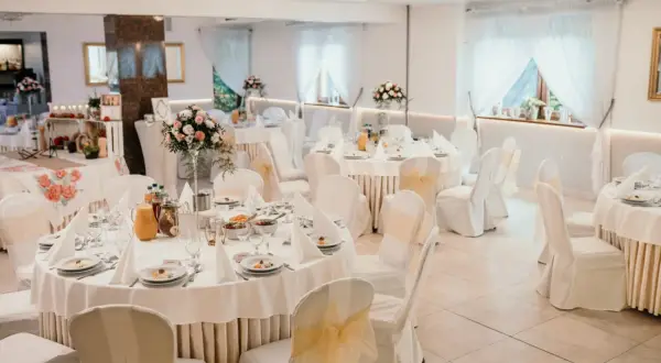 Elegancka sala weselna przygotowana na przyjęcie dla 100 osób. Stoły nakryte białymi obrusami, ozdobione kwiatami i zastawą. Krzesła w pokrowcach z kokardami.