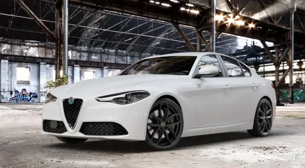 Biała Alfa Romeo Giulia Veloce z czarnymi felgami stoi w opuszczonej hali przemysłowej, gdzie światło przebija się przez dach.
